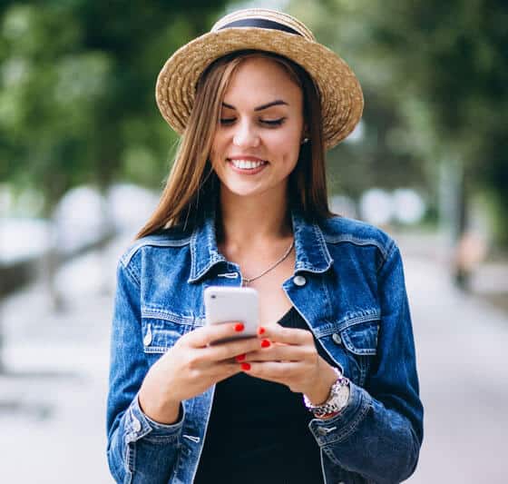 Hasır şapka ve kot ceket giyen genç bir kadın, arka planda ağaçlar ve bulanık bir patika ile açık havada akıllı telefonuna bakarken gülümsüyor ve muhtemelen CE belgesi veya TÜV Austria Türk akredite sertifika güncellemelerini okuyor.